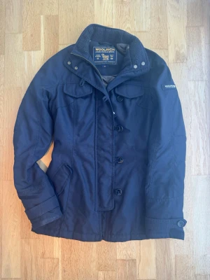 Mörkblå jacka från Woolrich - Snygg mörkblå jacka från Woolrich med klassisk krage, knappar och dragkedja framtill. Jackan har fickor med lock och en diskret logga på ärmen. Perfekt för kyligare dagar och har en stilren look.