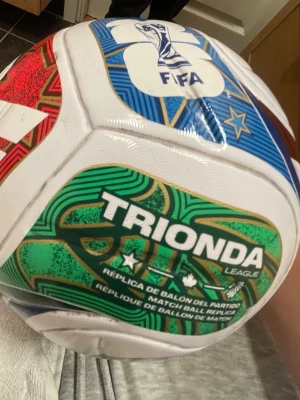 Vit och grön fotboll Trionda League - Säljer en snygg fotboll från Trionda League i vitt med gröna, blå och röda detaljer. Bollen har FIFA-logga och coola mönster, perfekt för matcher eller träning. Passar dig som vill ha en boll med unik design. Kan tvättas bättre om behövs . Har använt bollen 2 gånger säljer pga har för många och den använde jag knappt