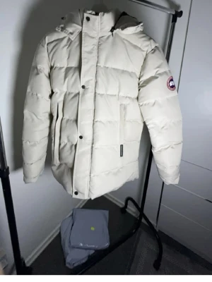 Vit dunjacka från Canada Goose - Snygg vit dunjacka från Canada Goose med huva och klassisk patch på ärmen. Jackan har stora fickor framtill, dragkedja och tryckknappar. Perfekt för kalla vinterdagar och riktigt varm tack vare dunfyllningen.