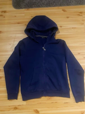 Mörkblå zip hoodie med fickor - En enkel och stilren mörkblå hoodie med dragkedja framtill och två stora fickor. Hoodien har en klassisk huva och ribbade muddar vid ärmar och nederkant. Perfekt för en avslappnad look och passar till det mesta.