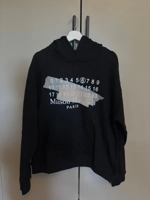 Svart Maison Margiela hoodie - Snygg svart hoodie från Maison Margiela med ikoniskt tryck framtill i vitt och grått. Oversized passform med huva och långa ärmar. Tillverkad i mjukt bomullsmaterial som känns skönt mot huden. Perfekt för en trendig och avslappnad stil..                                Använd någon gång endast! Har ej kvitto!  
