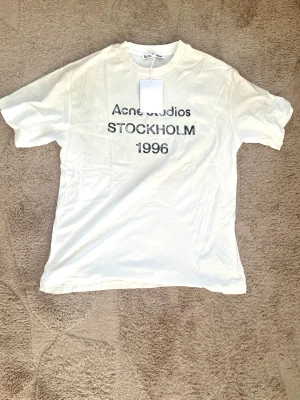 Vit Acne Studios t-shirt Stockholm - Vit t-shirt från Acne Studios med svart text 'STOCKHOLM 1996' på bröstet. Klassisk passform och rund halsringning. Perfekt basic-plagg med stilren design och mjukt material.