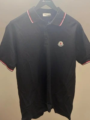 Moncler Piké L - Klassisk svart pikétröja från Moncler med korta ärmar och vit-röda detaljer på krage och ärmslut. Tröjan har Moncler-logga på bröstet och knäppning framtill. Perfekt till en clean och sportig stil. Storlek L helt ny oanvänd 