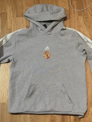 Grå Manchester United hoodie Adidas - Snygg grå hoodie från Adidas med Manchester United-logga på bröstet och klassiska vita ränder på ärmarna. Tröjan har huva och magficka, perfekt för dig som gillar sportig stil och vill visa vilket lag du hejar på.