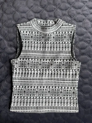 Svartvit mönstrad ärmlös topp - Snygg ärmlös topp från DIVIDED i svartvitt mönster. Toppen har hög hals och ett coolt grafiskt tryck som ger en trendig vibe. Perfekt att styla med jeans eller kjol för en modern look.