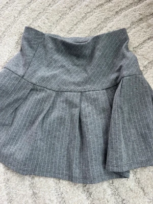Grå randig kjol från H&M - Säljer en grå kortkjol från H&M med smala vita ränder. Kjolen har en markerad midja och lätt utställd form med fina veck.  Storlek: 122/128