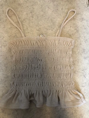 Beige smockad linnetopp med volang - Supersöt beige linnetopp med smockad resår över hela toppen och volangkant nedtill. Tunna axelband och mjukt material som känns skönt mot huden. Perfekt för varma dagar och enkel att styla med jeans eller kjol.
