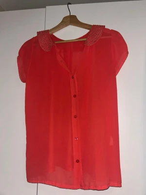 Röd transparent blus med nitar - Snygg röd blus i transparent material med korta ärmar och knäppning framtill. Kragen är dekorerad med små silvriga nitar som ger en cool touch. Perfekt att styla lager på lager eller med ett linne under.