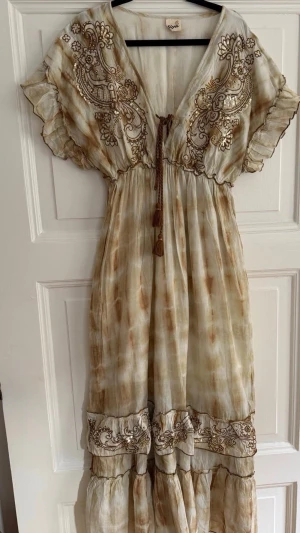 Beige långklänning med broderi - Bohemisk långklänning i beige och bruna toner med broderade detaljer och volangkant. Klänningen har v-ringning, korta ärmar och knytband med tofsar framtill. Tillverkad i ett tunt, luftigt material som passar perfekt för varma dagar.