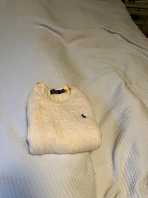 Vit kabelstickad tröja från Polo Ralph Lauren - Kabelstickad vit tröja från Polo Ralph Lauren med rund halsringning och den klassiska broderade loggan på bröstet. Tröjan har långa ärmar och är gjord i ett mjukt material som passar perfekt till jeans
