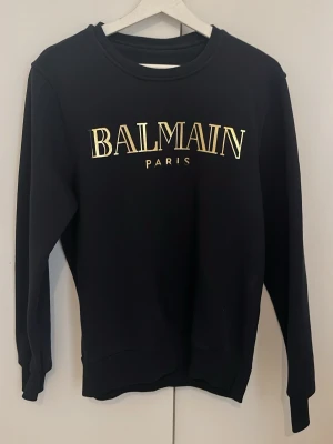 Svart tröja från Balmain - Snygg svart hoodie från Balmain Paris med guldfärgad logga på bröstet. Klassisk rak passform, rund halsringning och ribbade muddar vid ärmslut och nederkant. Perfekt för dig som vill ha en stilren och trendig look.