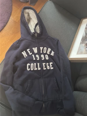 Marinblå hoodie med teddyfoder - Mysig marinblå hoodie med dragkedja och vit text 'NEW YORK 1990 COLLEGE' på bröstet. Utrustad med huva fodrad i vitt teddy-material och två fickor framtill. Perfekt för chill dagar och streetwear-vibe.