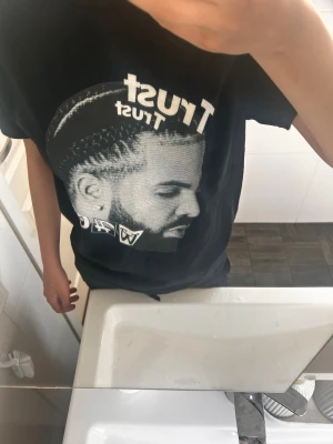 Drake t -shirt - Cool svart t-shirt med stort grafiskt tryck av ett ansikte och texten 'Trust' på framsidan. T-shirten har rund hals och korta ärmar, perfekt för en avslappnad streetwear-look.