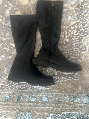 Svarta höga fashion stövlar - Säljer ett par svarta höga stövlar med dragkedja på sidan och chunky gummisula. Stövlarna är i ett slätt material och har en stilren, enkel design som passar till många olika outfits. Perfekta för dig som vill ha både stil och funktion under höst och vinter.