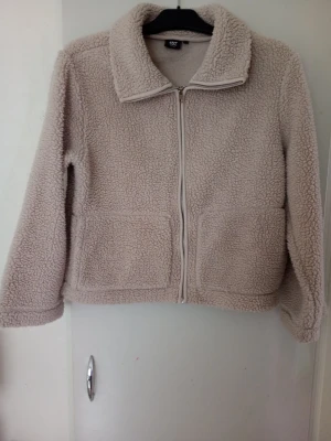 Beige teddyjacka med dragkedja - Mysig beige teddyjacka med hög krage och hel dragkedja framtill. Jackan har två stora fickor fram och är gjord i fluffigt teddy-material som håller dig varm. Perfekt för kyliga dagar och ger en avslappnad, trendig look.