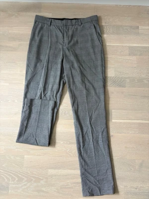 Grå rutiga kostymbyxor H&M Slim Fit - Snygga grå kostymbyxor från H&M i slim fit-modell med diskret rutmönster. Byxorna har hällor för bälte, snedställda fickor och klassisk pressveck. Perfekta för en stilren och modern look.