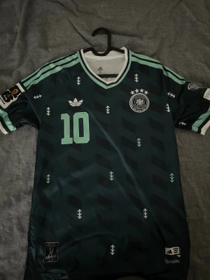 Tysklands VM 2026 Musiala - Snygg mörkgrön Tyskland fotbollströja från Adidas med Musiala och nummer 10 på ryggen. Tröjan har mintgröna detaljer, V-ringning och vita ränder på axlarna. Officiella märken och Climacool-material för bästa komfort under match eller träning.