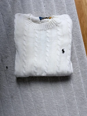 Stickad Ralph Lauren Tröja - Klassisk vit kabelstickad tröja från Ralph Lauren med rund halsringning och broderad logga på bröstet. Tröjan har ribbade muddar och är gjord i mjukt material som passar perfekt till många olika outfits.