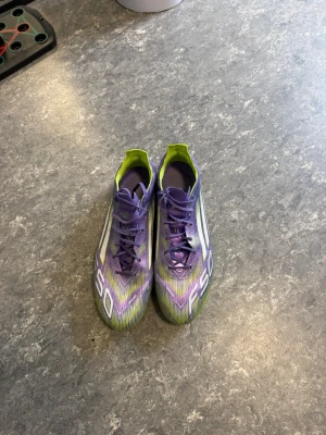 Lila Adidas F50 elite fotbollsskor - Säljer ett par Adidas F50 fotbollsskor i lila, vitt och neongrönt. Skorna har snörning, låg profil och tydliga vita stripes på sidan. Snygg gradientdesign och F50-tryck på ovansidan. Perfekta för dig som vill sticka ut på planen. Vi kan även diskutera om priset och ni får även väskan med.