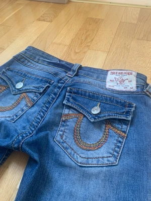 Blå bootcut jeans från True Religion - Snygga blå bootcut jeans från True Religion med ikoniska broderade bakfickor och silverfärgade knappar. Jeansen har klassisk femficksdesign och är tillverkade i denim med lätt tvättad look. Perfekta för dig som gillar en avslappnad men trendig stil.