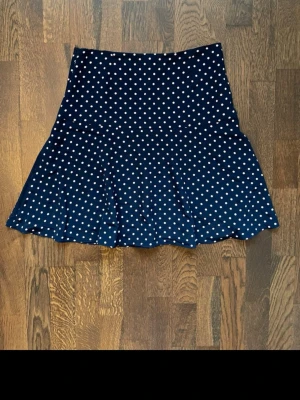 Mörkblå prickig kjol från Ralph Lauren - Snygg mörkblå kjol från Lauren Ralph Lauren med vita prickar och lätt utställd form. Kjolen är gjord i mjuk polyester och har en klassisk, tidlös look som passar perfekt till många olika stilar.