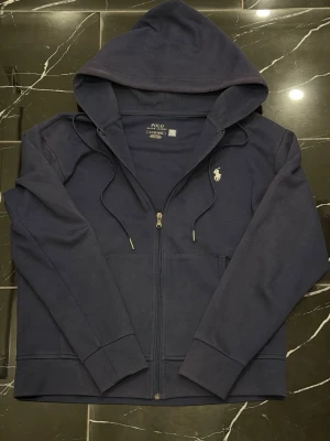 Mörkblå Zip Hoodie från Polo Ralph Lauren - Mörkblå hoodie med dragkedja från Polo Ralph Lauren. Klassisk design med huva, dragsnören och kängurufickor. Ikoniska vita logon broderad på bröstet. Tillverkad i mjukt bomullsmaterial för extra komfort. Tröjan är knappt använd och i ett jättebra skick!!