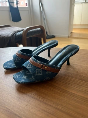 Blå denim-pumps med brunt band - Snygga pumps i blått denimtyg med klassiskt monogrammönster och brunt läderband över foten. Skorna har öppen tå och smal klack för en trendig look. Perfekta för dig som vill sticka ut med en unik och cool stil.