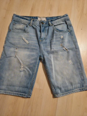 Ljusblå slitna jeansshorts - Säljer ett par ljusblå jeansshorts med slitna detaljer och råa kanter. Shortsen har fem fickor, bälteshällor och knappgylf. Perfekta för dig som gillar en avslappnad och trendig stil.