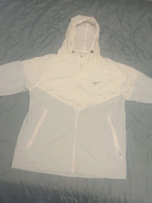Nike Miller - Snygg vit vindjacka från Nike med dragkedja framtill, två fickor med dragkedja och justerbar huva. Jackan har en sportig look med diskret Nike-logga på bröstet och är tillverkad i lätt material som passar perfekt för blåsiga dagar.