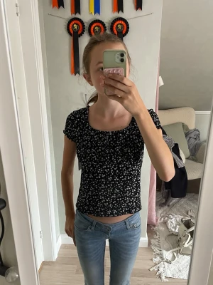 Svart blommig croppad topp - Snygg svart croppad topp med små vita blommor och korta ärmar. Toppen har en rundad halsringning och sitter tight. Perfekt till jeans eller kjol för en trendig look.