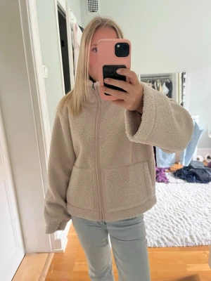 Beige teddyjacka med dragkedja - Mysig beige teddyjacka med hög krage och dragkedja framtill. Jackan har två stora fickor och vida ärmar för en avslappnad look. Perfekt för att hålla värmen under kyliga dagar och ger en trendig vibe till din outfit.