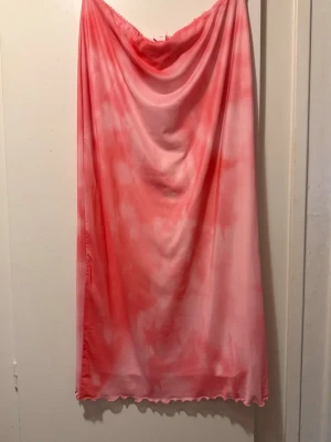 Rosa-röd långkjol - Snygg långkjol med tie-dye effekt i rosa och rött. Kjolen har vågig kant nertill och ett mjukt, glansigt material som faller fint. Perfekt för dig som vill sticka ut med en färgstark look.