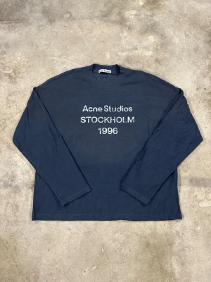 Mörkblå långärmad t-shirt Acne Studios - Snygg mörkblå långärmad t-shirt från Acne Studios med tryck 'STOCKHOLM 1996' på bröstet. Klassisk passform och rund halsringning, perfekt för en stilren och avslappnad look. Storlek M, se bilder för mått 