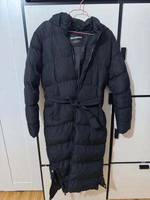 Jacka med knyt  - Snygg lång svart dunjacka från Threadbare med huva och bälte i midjan. Jackan har dragkedja framtill, två fickor och slitsar nedtill med knappar. Perfekt för kalla vinterdagar och ger en stilren look. Inga defekter. Från asos. Använd 1 gång