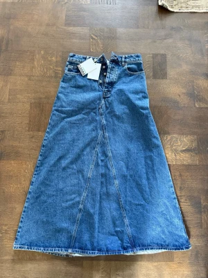 Blå lång jeanskjol från Ganni - Säljer en blå lång jeanskjol från Ganni med klassisk femficksmodell och synliga knappar framtill. Kjolen har en A-linjeformad siluett och är tillverkad i kraftigt denimtyg. Perfekt för dig som gillar en avslappnad men trendig stil.