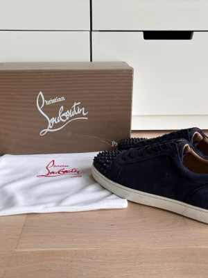  Christian Louboutin sneakers - Snygga sneakers från Christian Louboutin med klassiska röda sulor och svarta nitar på tån. Skorna har en låg profil och är tillverkade i mocka med vita gummisulor. Perfekta för dig som vill sticka ut med exklusiv design och edgy detaljer.