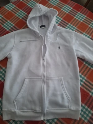 Vit hoodie med dragkedja från Polo Ralph Lauren - Säljer en stilren vit hoodie från Polo Ralph Lauren med dragkedja framtill och klassisk huva. Tröjan har två stora fickor fram och den ikoniska lilla loggan broderad på bröstet. Perfekt för en clean och avslappnad look.