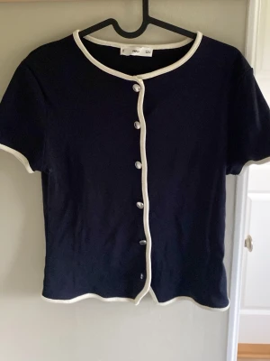 Mörkblå kofta med vita kanter MNG - Kortärmad mörkblå kofta från MNG med vita kontrastkanter och knappar framtill. Snygg, enkel och stilren design som passar till mycket. Materialet känns mjukt och stretchigt, perfekt för lager på lager eller sommaroutfits.