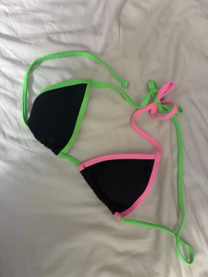 Svart bikiniöverdel med neonkanter - Snygg trekantsbikini i svart med neonrosa och neongröna kanter. Klassisk modell med knyt i nacke och rygg, perfekt för sommaren och strandhäng. Färgstarka detaljer som verkligen poppar!