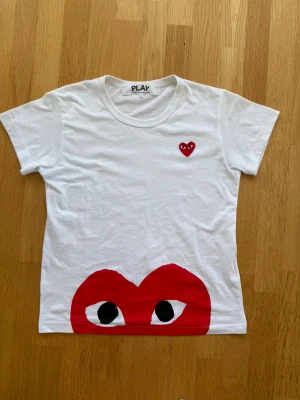 Vit Comme des Garçons PLAY t-shirt - Säljer en vit t-shirt från Comme des Garçons PLAY med det ikoniska röda hjärtat med ögon på bröstet och ett stort hjärtmotiv längst ner. Klassisk rund hals och korta ärmar. Perfekt för dig som gillar streetwear och vill sticka ut.