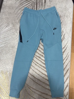 Blå mjukisbyxor från Nike - Snygga ljusblå mjukisbyxor från Nike med svarta detaljer vid fickorna och justerbar dragsko i midjan. Byxorna har ribbade muddar vid bensluten och en modern, avsmalnande passform. Perfekta för chill eller träning.