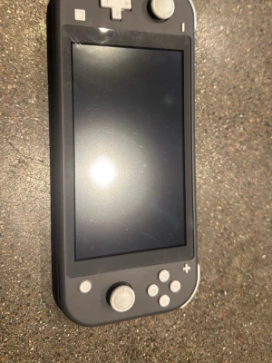 Grå Nintendo Switch Lite konsol - Säljer en grå Nintendo Switch Lite, en smidig och portabel spelkonsol med inbyggda knappar och styrspakar. Perfekt för gaming på språng och har en matt finish i plast. Skärmen är centrerad och omgiven av ljusgrå knappar och styrkors. Du får med dig lådan o ett minneskort 150gb 