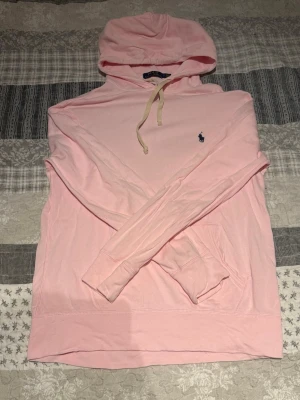 Ljusrosa hoodie med huva från Ralph Lauren - Säljer en ljusrosa hoodie med huva och dragsnören. Tröjan har en klassisk känguruficka framtill och en liten broderad logga på bröstet. Tillverkad i mjukt bomullstyg och har långa ärmar. Perfekt för en avslappnad och stilren look.