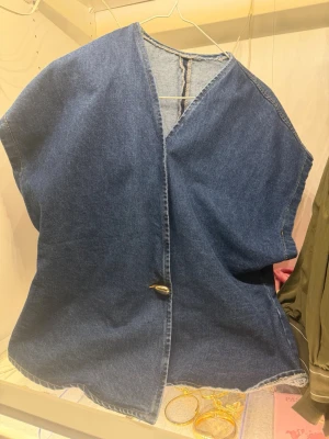 Oversized jeansväst i denim - Säljer en oversized jeansväst i klassisk blå denim med råa kanter och en enkel knapp framtill. Västen har en avslappnad passform och är perfekt för att ge din outfit en cool och trendig touch.