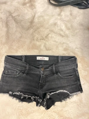 Svarta  jeansshorts med fransig kant - Snygga svarta  jeansshorts från Hollister med fransig, rå kant nertill och klassisk femficksdesign. Shortsen har låg midja och slitna detaljer för en avslappnad look. Perfekta till sommaren och lätta att matcha med olika toppar.