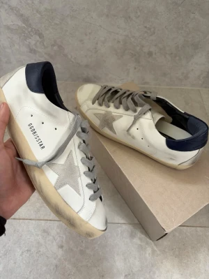 Golden Goose vita sneakers med stjärna - Golden Goose sneakers i vitt läder med grå mockastjärna på sidan, grå skosnören och mörkblå häl. Klassisk låg modell med slitna detaljer och beige sula för en trendig look. Perfekta för dig som gillar streetstyle och unika detaljer.