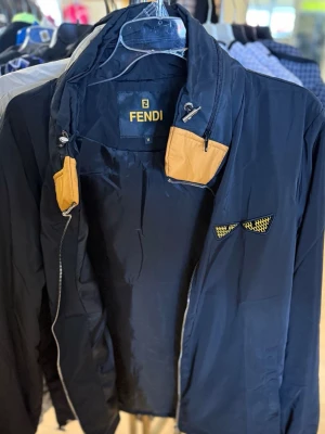 vindjacka från Fendi - Snygg vindjacka från Fendi med gulbruna detaljer vid kragen och en cool gulsvart detalj på bröstet. Jackan har dragkedja och är tillverkad i ett lätt, glansigt material som känns sportigt och modernt. Perfekt för dig som vill ha en stilren och trendig look.