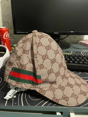 Beige keps med GG-mönster och ränder - Snygg beige keps med brunt GG-mönster över hela ytan och en grön-röd rand på sidan. Kepsen har böjd skärm och klassisk form, perfekt för att lyfta din streetwear-look. Materialet är tyg med tydlig struktur.