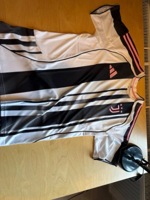 Juventus 25/26  - Snygg fotbollströja i vitt med svarta och rosa detaljer. Tröjan har korta ärmar, svarta ränder och rosa inslag på axlarna samt ett klubbmärke och tre stjärnor på bröstet. Tillverkad i ett lätt och ventilerande material, perfekt för träning eller match.