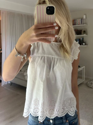 Vit blus med volangärm - Söt vit topp i bomull med broderade detaljer och volangärmar. Toppen har ett luftigt fall, rund halsringning och vackert broderad nederkant med vågig kant. Perfekt för sommarens alla tillfällen och ger en romantisk vibe.